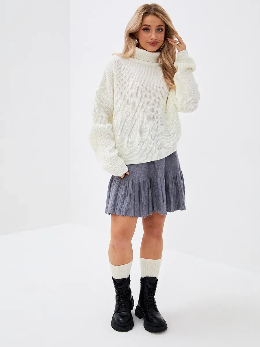 Model wearing the Lyra Sweater by Lauven, oversized knit with high neck in ivory, paired with a pleated skirt.