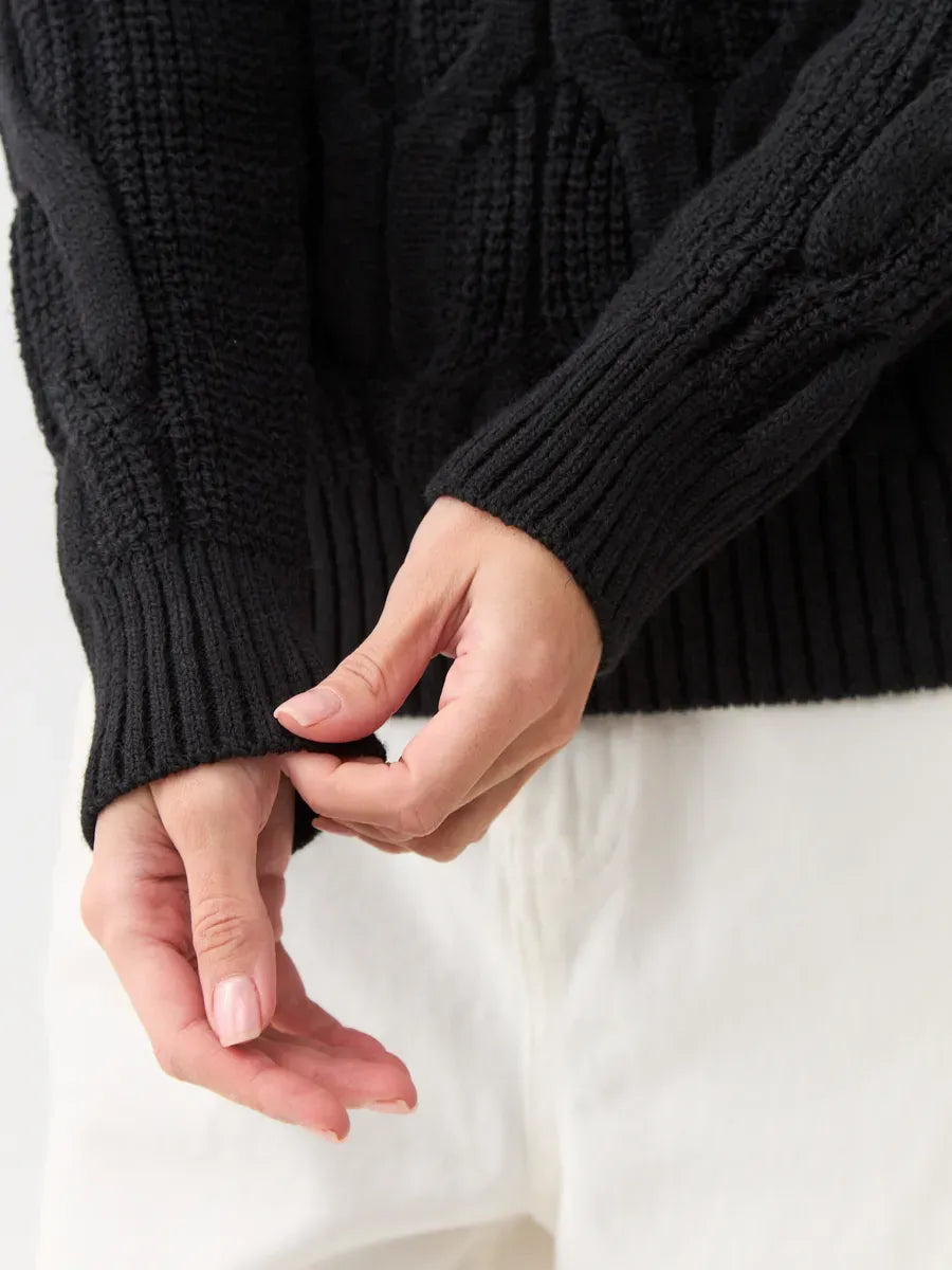 Close-up of hands adjusting sleeves of the Noir Knitwear by Lauven, a wool-blend turtleneck sweater in black.