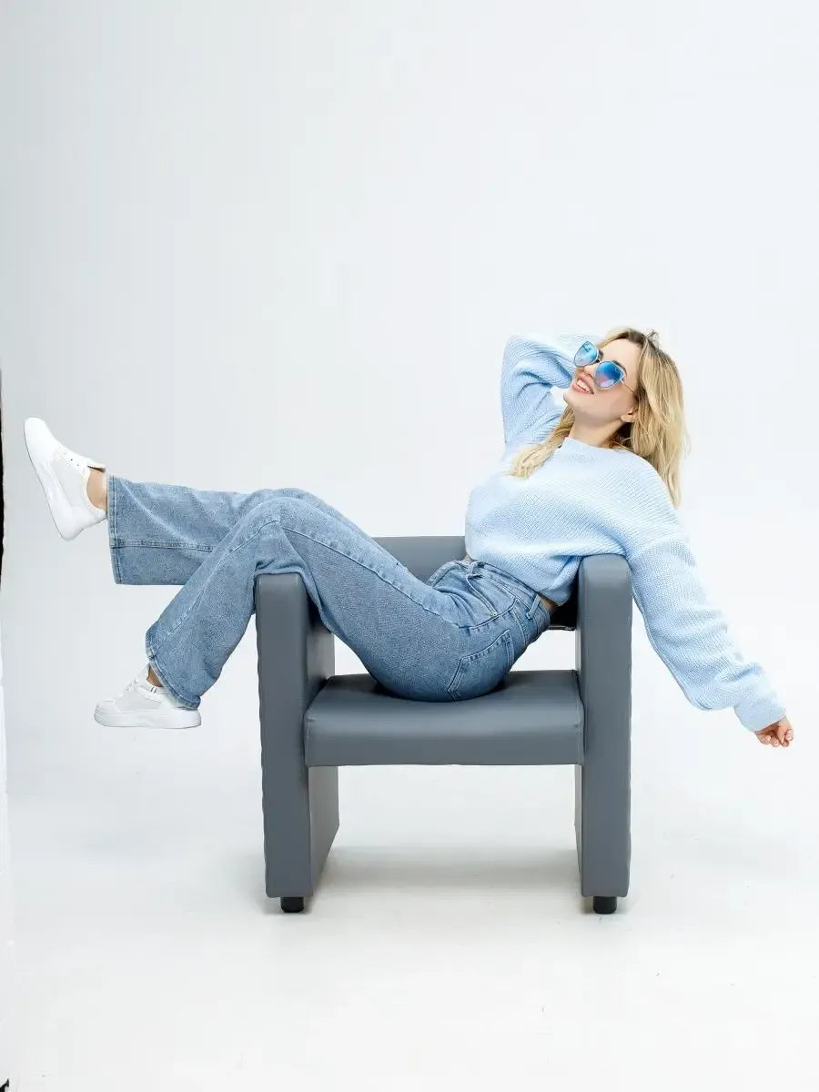 Model in a Skylar Pullover by Lauven, relaxed in a gray chair, showcasing casual spring style with light blue hues.