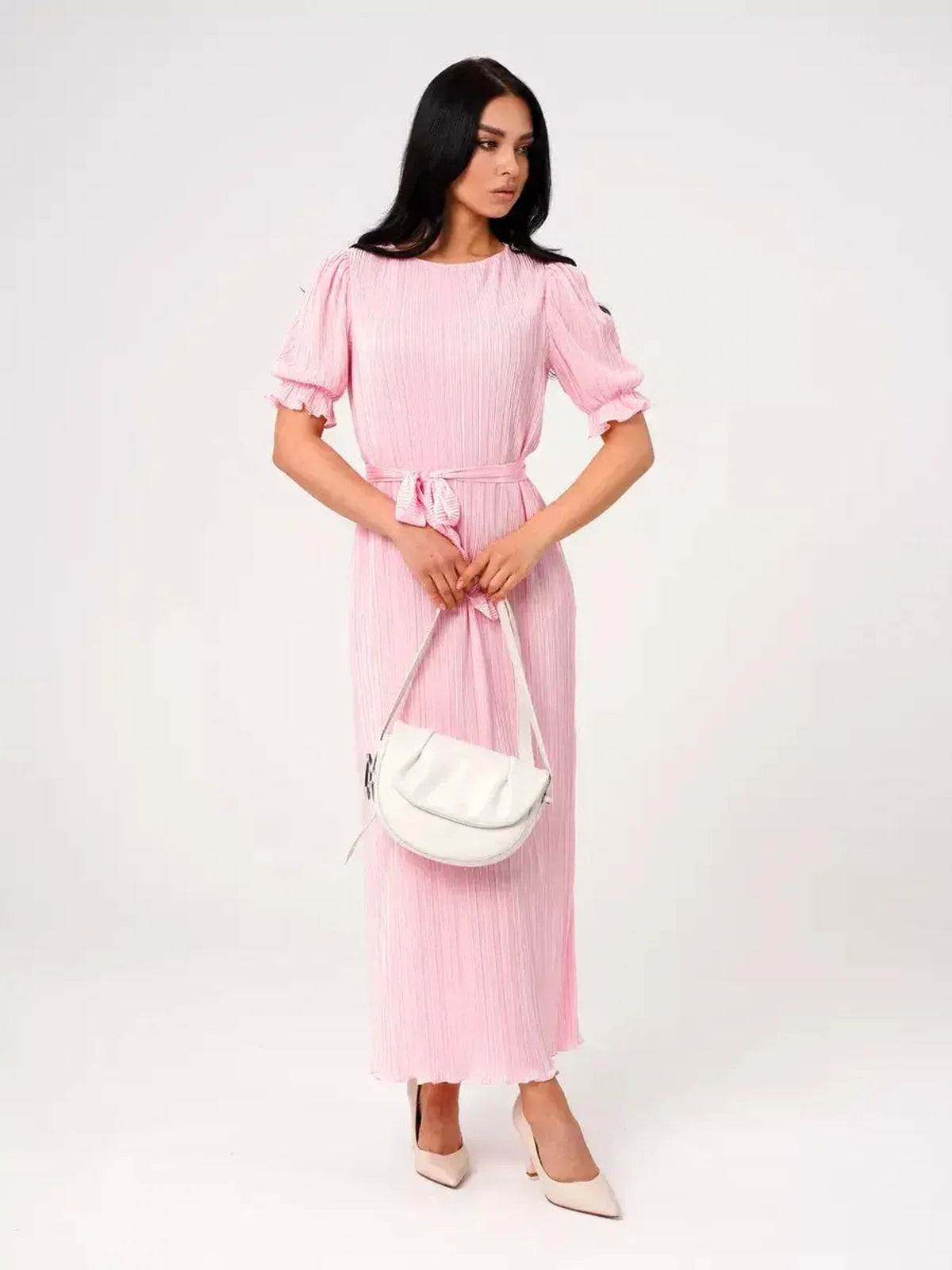 Model wearing the Candy Maxi Dress, a pink pleated dress with puff sleeves and a tie waist, paired with a stylish handbag.