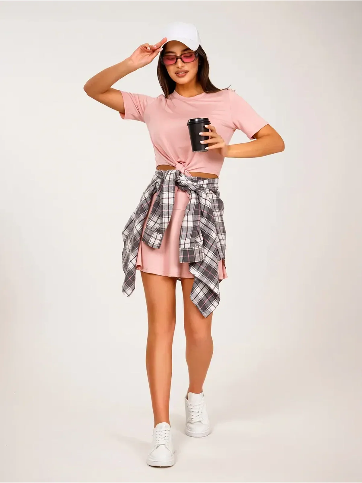 Woman in pink cropped top and mini skirt, wearing a plaid shirt and cap, holding a drink.