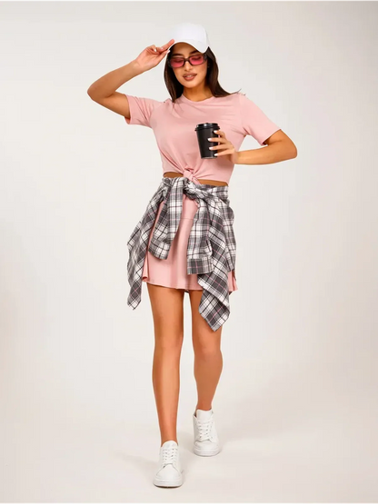 Woman in pink cropped top and mini skirt, wearing a plaid shirt and cap, holding a drink.