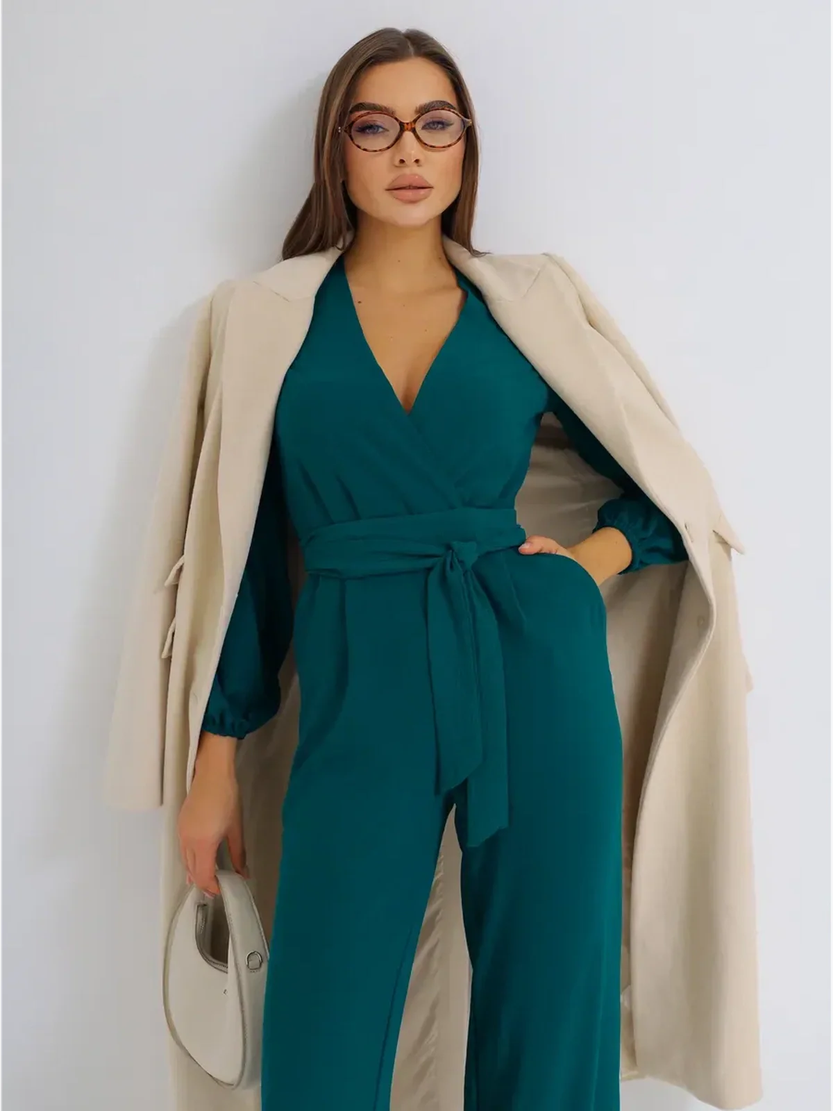 Model wearing the Zoe Jumpsuit with a belted waist and long sleeves, styled with a beige coat and glasses.