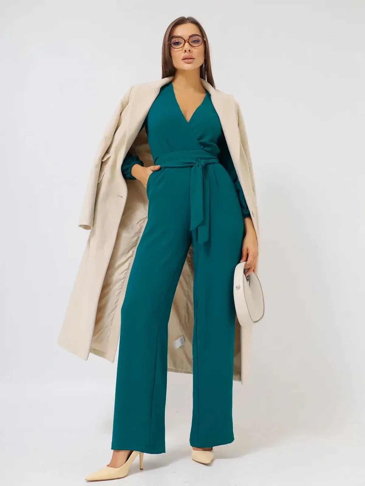 Model wearing a Zoe Jumpsuit in teal with wide-leg trousers, a self-tie belt, and a stylish coat, showcasing chic fashion.